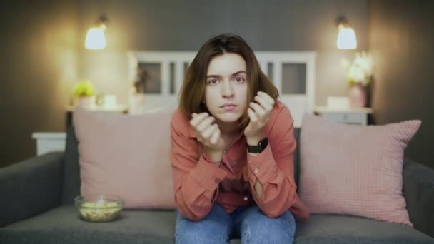 Joven mujer entusiasta sentada en el sofá, comiendo palomitas de maíz y viendo la televisión — Vídeo de stock