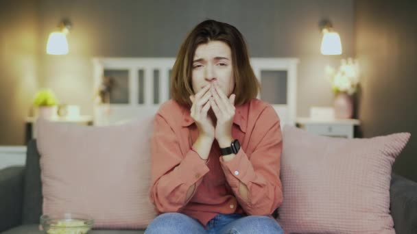 Szomorú fiatal nő ül a kanapén, popcornt eszik, melodrámát néz és sír. — Stock videók