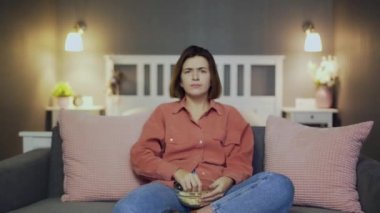 Tatminsiz ve hayal kırıklığına uğramış bir kadın kanepede oturur, televizyon izler ve patlamış mısır fırlatır.