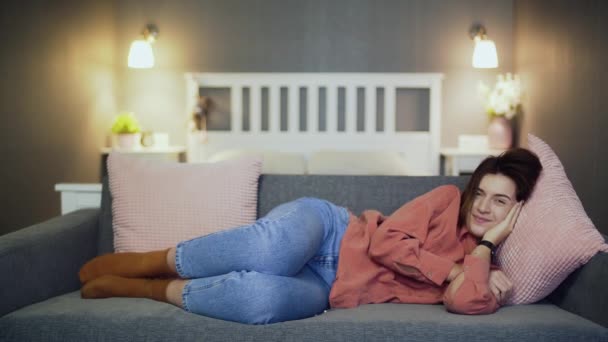 Счастливая молодая женщина лежит на диване, смотрит комедийный фильм и смеется — стоковое видео