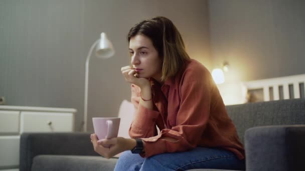 Junge enttäuschte Frau sitzt mit Tasse Kaffee auf dem Sofa und schaut fern — Stockvideo