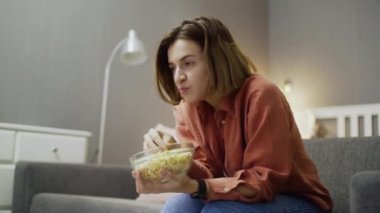 Kanepede oturan, patlamış mısır yiyen ve televizyon izleyen genç bir kadın.