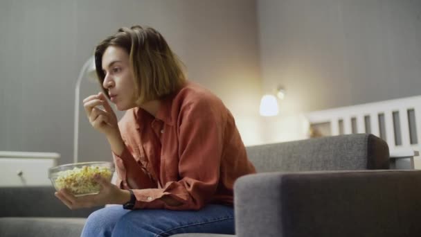 Νεαρή ενθουσιώδης γυναίκα κάθεται στον καναπέ, τρώει ποπ κορν και βλέπει τηλεόραση — Αρχείο Βίντεο