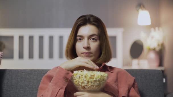 Primer plano de una joven sentada en el sofá, comiendo palomitas de maíz y viendo la televisión — Vídeos de Stock