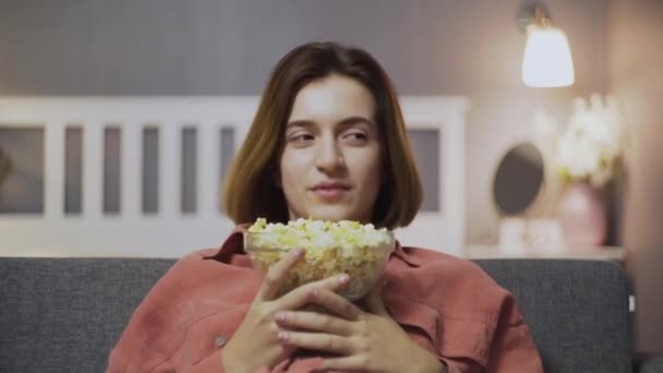 Primer plano de la joven feliz sentada en el sofá, comiendo palomitas de maíz, viendo películas de comedia y riendo — Vídeo de stock