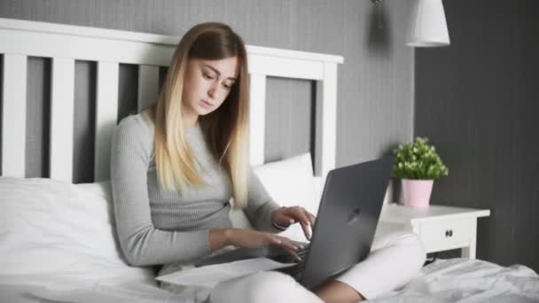 Mujer seria se sienta en la cama, utiliza el ordenador portátil y trabaja con documentos — Vídeo de stock