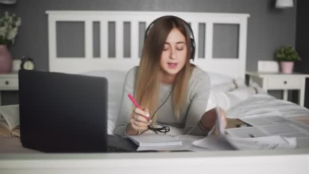 Молода жінка з навушниками лежить на ліжку, слухає музику і робить гарячі на ноутбуці — стокове відео
