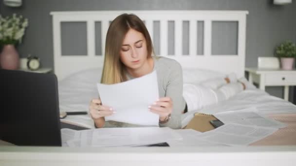 Een jonge vrouw werkt thuis. Vrouw ligt op het bed en werkt met documenten — Stockvideo