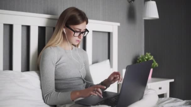Серьезная женщина в наушниках сидит на кровати, пользуется ноутбуком и работает с документами — стоковое видео