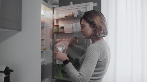 Közelkép a nő kinyitja a hűtőszekrény ajtaját a konyhában otthon, és vesz egy üveg tejet, majd önti a tejet az üveg — Stock videók