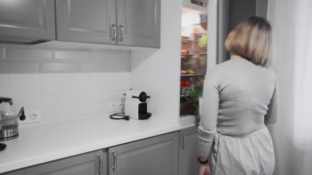 Mujer con dolor de cabeza o resaca abre la puerta del refrigerador en la cocina en casa y toma botella de agua — Vídeo de stock