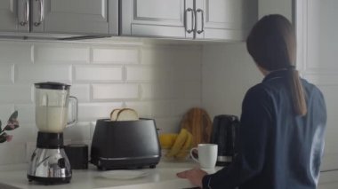 Siyah pijamalı genç bir kadın sabah kahvaltısını hazırlıyor. Kadın tost makinesini çalıştırır, elektrikli su ısıtıcısını çalıştırır ve mikserde kokteyl yapar.