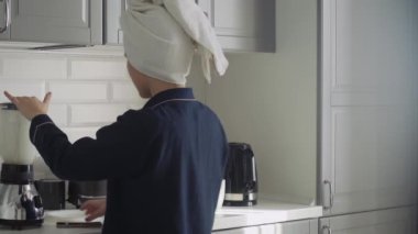 Siyah pijamalı ve kafasında havlu olan genç bir kadın sabah kahvaltısını hazırlıyor. Kadın mikserde kokteyl yapar ve tost makinesinden ekmek alır.