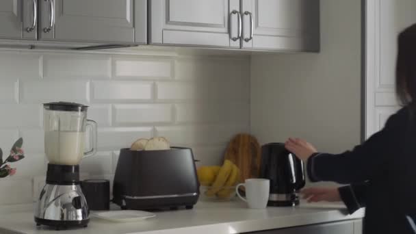 Mujer joven en pijama negro preparando el desayuno por la mañana. Mujer enciende la tostadora, Hervidor eléctrico y hace un cóctel en una licuadora — Vídeos de Stock