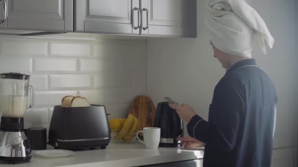 Mujer joven en pijama negro y toalla en la cabeza preparando el desayuno por la mañana. Mujer enciende la tostadora, Hervidor eléctrico y hace un cóctel en una licuadora — Vídeos de Stock