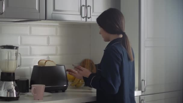 Jonge vrouw in zwarte pyjama bereidt ontbijt voor 's morgens. Vrouw met smartphone zet broodrooster aan en giet een warm water — Stockvideo
