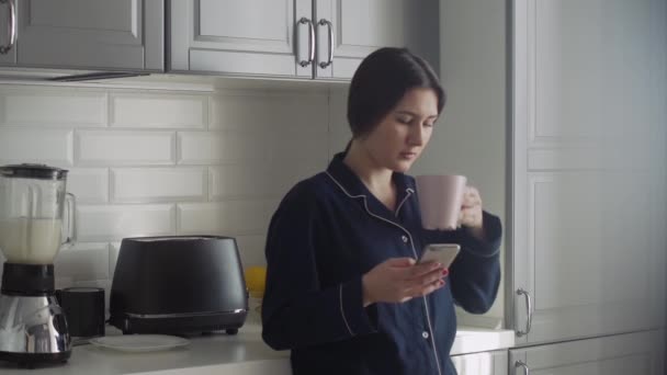 Wanita muda dengan piyama hitam menyiapkan sarapan di pagi hari. Wanita dengan smartphone dan secangkir kopi atau teh memeriksa pemanggang roti — Stok Video