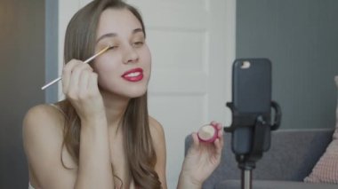 Güzellik blogcusu video kaydetmek için ön kamerada otururken güzellik kozmetiklerini sunuyor. Kadın göz kapağına fırça sürterek pudra sürüyor