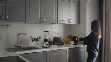 Siyah pijamalı genç bir kadın sabah kahvaltısını hazırlıyor. Kadın tost makinesini çalıştırır, elektrikli su ısıtıcısını çalıştırır ve mikserde kokteyl yapar.