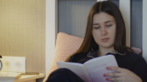 Jovem de pijama preto lê livro e usa telefone na cama em casa — Vídeo de Stock