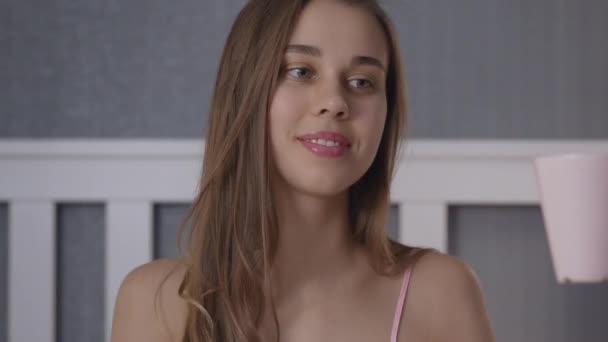 Ung vacker kvinna i rosa pyjamas sitter på sängen och dricker te eller kaffe — Stockvideo