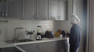 Siyah pijamalı ve kafasında havlu olan genç bir kadın sabah kahvaltısını hazırlıyor. Kadın tost makinesini çalıştırır, elektrikli su ısıtıcısını çalıştırır ve mikserde kokteyl yapar.