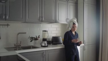 Siyah pijamalı ve kafasında havlu olan genç bir kadın sabah kahvaltısını hazırlıyor. Kadın akıllı telefon kullanıyor ve ekmek kızartma makinesinden ekmek alıyor.