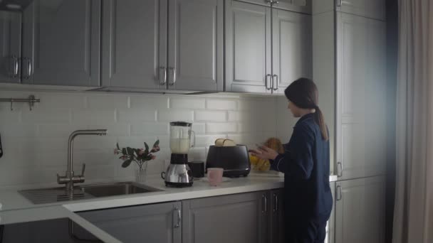 검은 잠옷을 입은 젊은 여성 이 아침 식사를 준비하고 있습니다. 스마트폰을 가진 여자가 토스터를 켜고 뜨거운 물을 붓는다 — 비디오