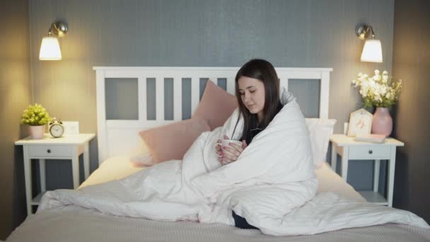 Jonge vrouw in zwarte pyjama verpakt in een deken drinkt koffie — Stockvideo