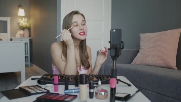 La blogueuse beauté présente des cosmétiques de beauté alors qu'elle est assise devant la caméra pour enregistrer des vidéos. Femme applique de la poudre avec pinceau sur la paupière — Video