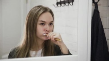 Banyoda diş fırçasıyla diş fırçalayan genç bir kadın. Aynadaki yansıma portresi.