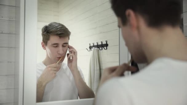 Young man talking on phone and holding wooden toothbrush brushing teeth looking in mirror — Stock Video
