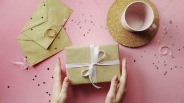 Kadın elleri beyaz kurdeleyle sarılı bir Noel hediyesini sallıyor. Tahta, pembe masa, düz uzanma.