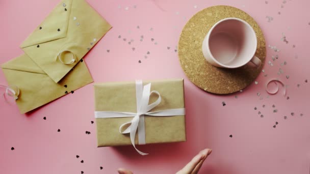 Manos de mujer sacudiendo un regalo de Navidad envuelto en papel artesanal con lazo blanco. Vista superior sobre tabla rosada de madera, posición plana — Vídeo de stock