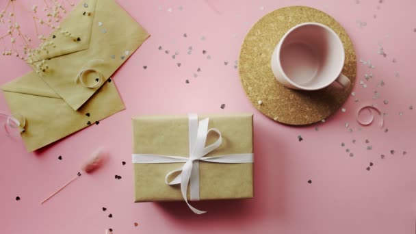 Manos de mujer tocando un regalo de Navidad envuelto en papel artesanal con lazo blanco. Vista superior sobre tabla rosada de madera, posición plana — Vídeos de Stock