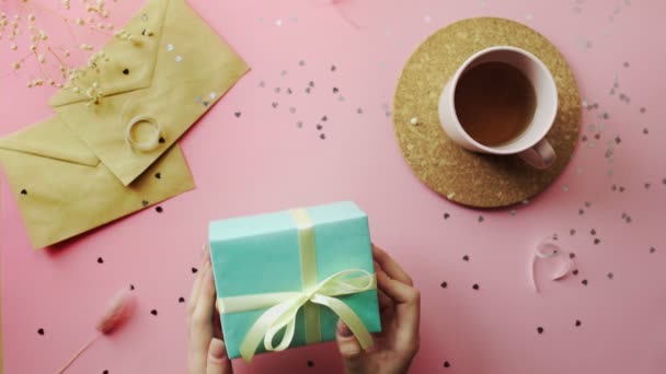 Vrouw handen schudden een kerstcadeau verpakt in een groen papier met gele strik. Bovenaanzicht op hout roze tafel, plat gelegd — Stockvideo