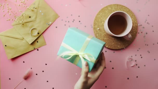 Manos de mujer agitando un regalo de Navidad envuelto en un papel verde con lazo amarillo. Vista superior sobre tabla rosada de madera, posición plana — Vídeos de Stock