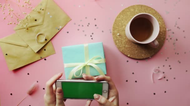 Mani donna scattare una foto di un regalo di Natale avvolto in una carta verde con fiocco giallo. Vista dall'alto su tavolo in legno rosa, disposizione piatta — Video Stock