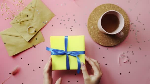 Woman hands putting a Christmas gift wrapped in a yellow paper with blue bow on top view on wood pink table, flat lay — Stock Video