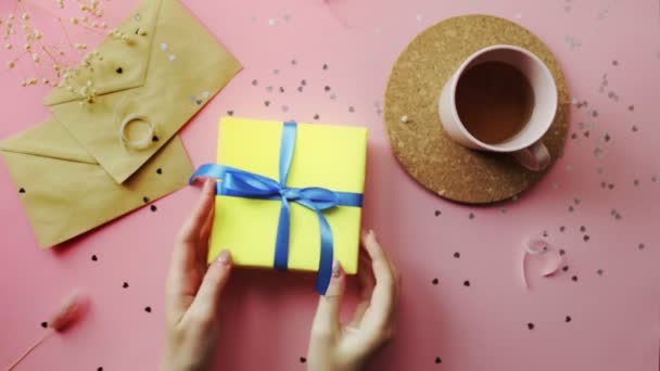 Manos de mujer tratando de abrir un regalo de Navidad envuelto en un papel amarillo con lazo azul. Vista superior sobre tabla rosada de madera, posición plana — Vídeos de Stock