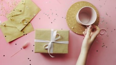 Kadın eli, beyaz kurdeleyle sarılmış bir Noel hediyesini inceliyor. Tahta, pembe masa, düz uzanma.