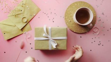 Kadın eli, beyaz kurdeleyle sarılı bir Noel hediyesini bağlar. Tahta, pembe masa, düz uzanma.