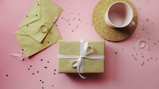 Woman hands shaking a Christmas gift wrapped in craft paper with white bow. Top view on wood pink table, flat lay — Stock Video