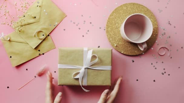 Mains de femme prenant et remettant un cadeau de Noël enveloppé dans du papier artisanal avec arc blanc. Vue du dessus sur table rose bois, plan plat — Video