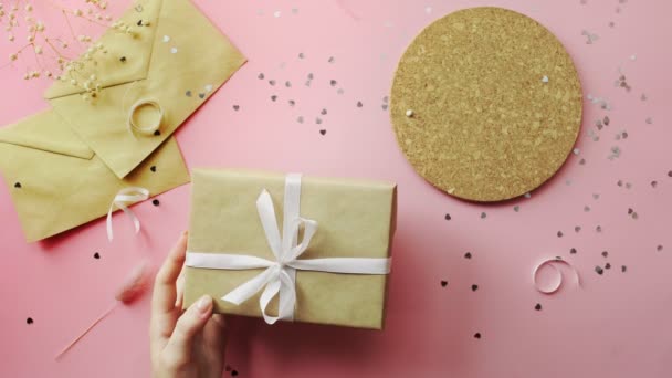 Las manos femeninas atan un regalo de Navidad envuelto en papel artesanal con lazo blanco. Vista superior sobre tabla rosada de madera, posición plana — Vídeo de stock