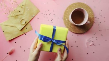 Kadın eli, mavi kurdeleli sarı kağıda sarılı bir Noel hediyesini bağlıyor. Tahta, pembe masa, düz uzanma.