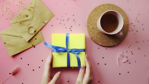 Mains de femme prenant et remettant un cadeau de Noël enveloppé dans du papier jaune avec arc bleu. Vue du dessus sur table rose bois, plan plat — Video