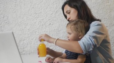 Genç anne bebeğine şişeden meyve suyu veriyor ve defter üzerinde çalışıyor.