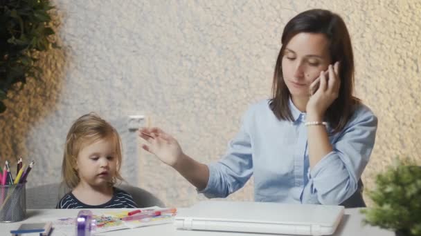 A fiatal anya simogatja a babát és telefonon beszél. Egy szülési szabadságon lévő nő otthonról dolgozik, fontos telefonhívásokat bonyolít, fogja a babát, a laptop előtt ül.. — Stock videók