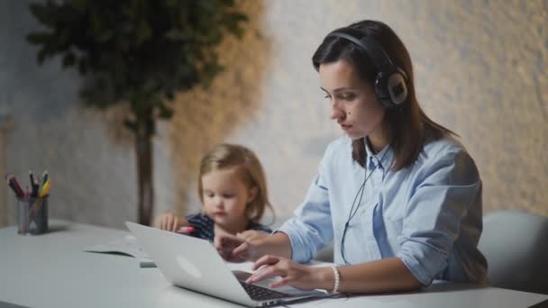 Mujer joven con auriculares trabajando en casa mientras su pequeño bebé está pintando. Madre usando notebook para el surf, freelance o educación — Vídeos de Stock